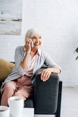 Kanepede oturmuş gülümseyen ve akıllı telefondan konuşan son sınıf öğrencisi kadın. 