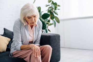 senior woman having knee Arthritis and sitting on sofa in apartment  clipart