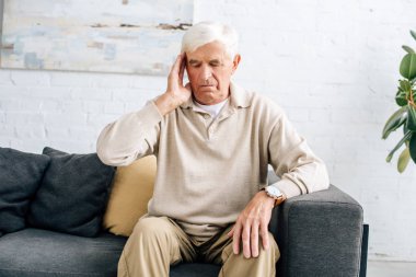 senior man sitting on sofa and having headache in apartment  clipart