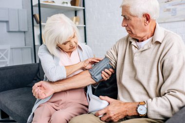 wife wearing blood pressure monitor on husband in apartment  clipart