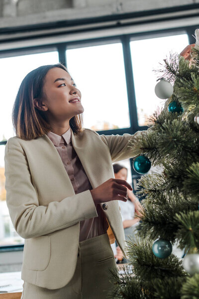 happy asian businesswoman looking at decorated christmas tree in office  