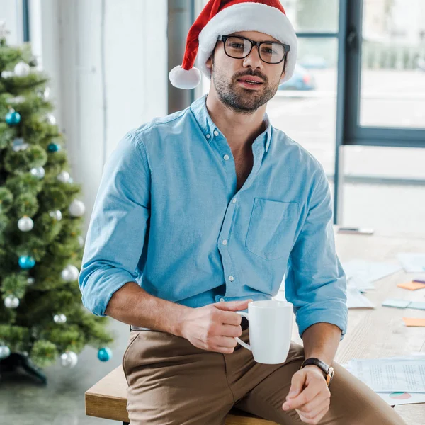 stock image selective focus of happy bearded man in santa hat and glasses holding cup with coffee near christmas tree in office 