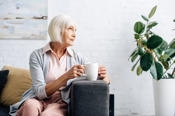 Starsza Kobieta Siedzi Kanapie Trzyma Filiżankę Herbaty Mieszkaniu — Zdjęcie stockowe