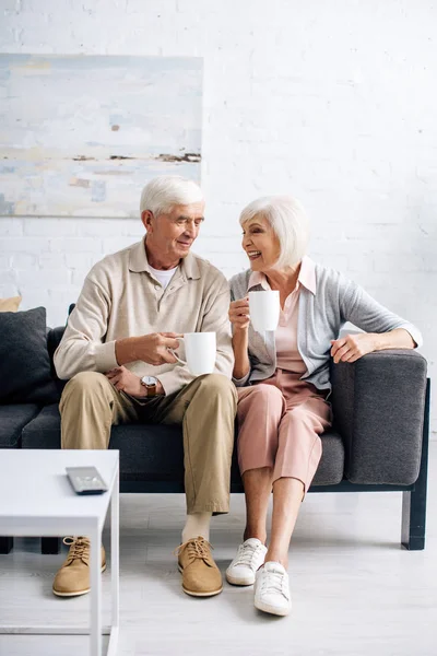 Lächelndes Ehepaar Hält Tassen Der Hand Und Sitzt Auf Sofa — Stockfoto