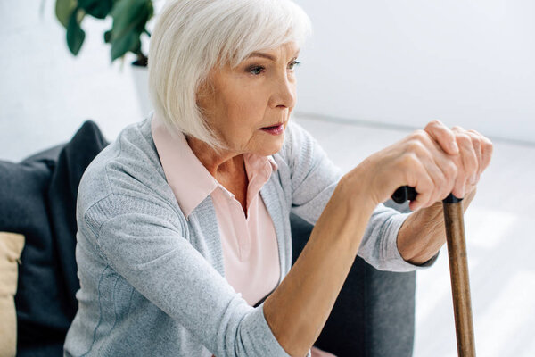 senior woman with wooden cane sitting on sofa in apartment 