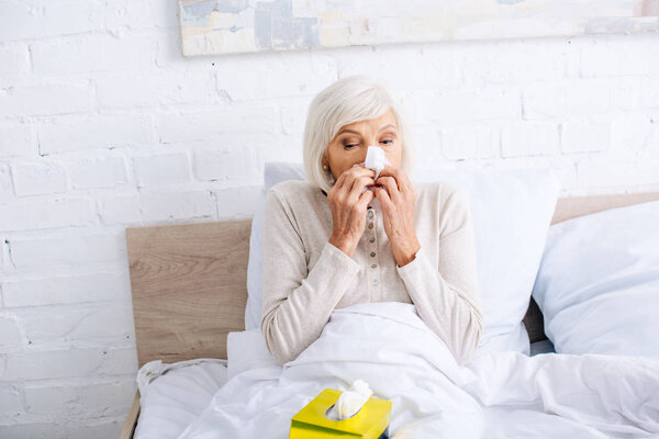 ill senior woman sneezing and holding napkin in bed 