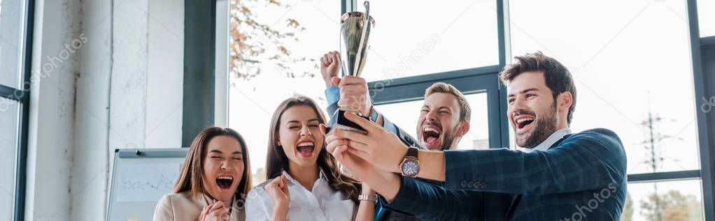 panoramic shot of excited multicultural businesswomen and businessmen looking at trophy in office 