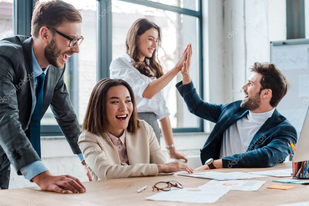 selective focus of happy businessman in glasses smiling near multicultural coworkers giving high five in office 