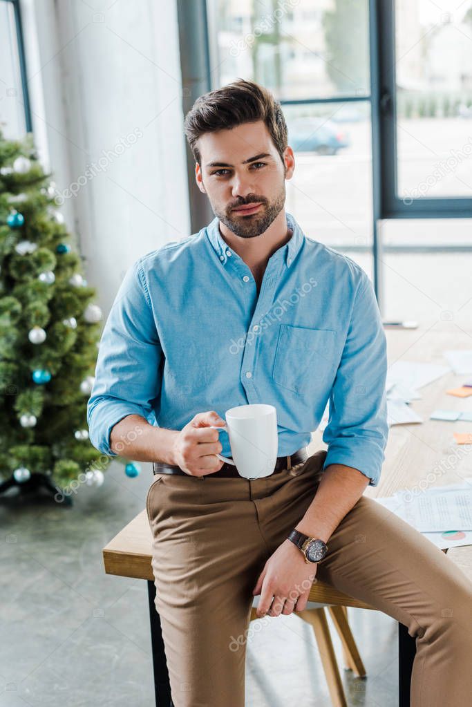 selective focus of bearded man holding cup near christmas tree in office 