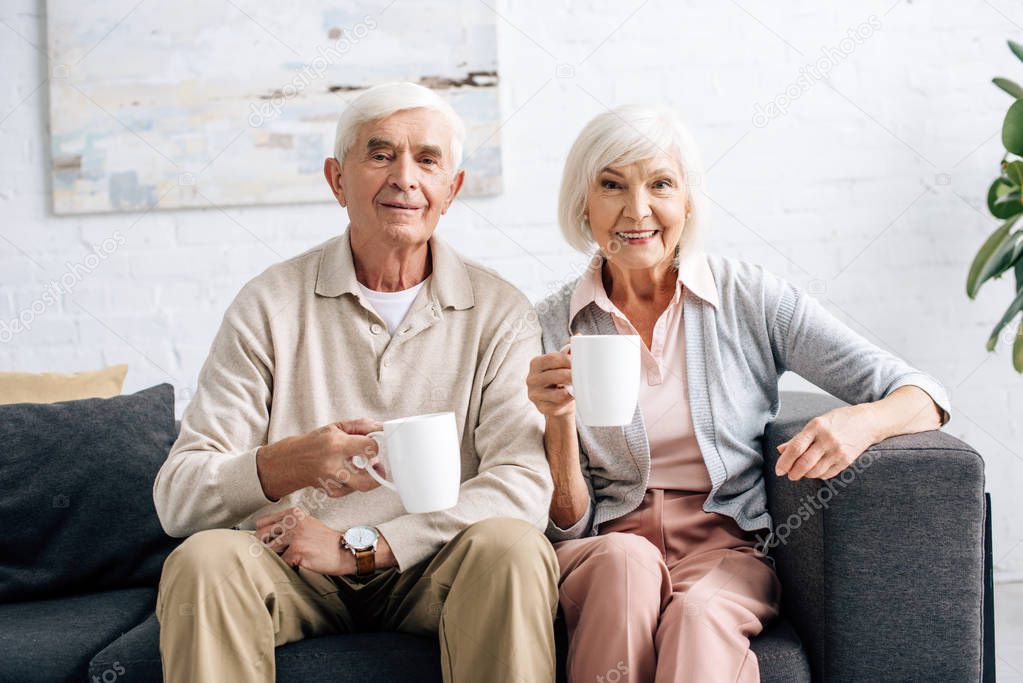 smiling husband and wife holding cups and sitting on sofa in apartment 