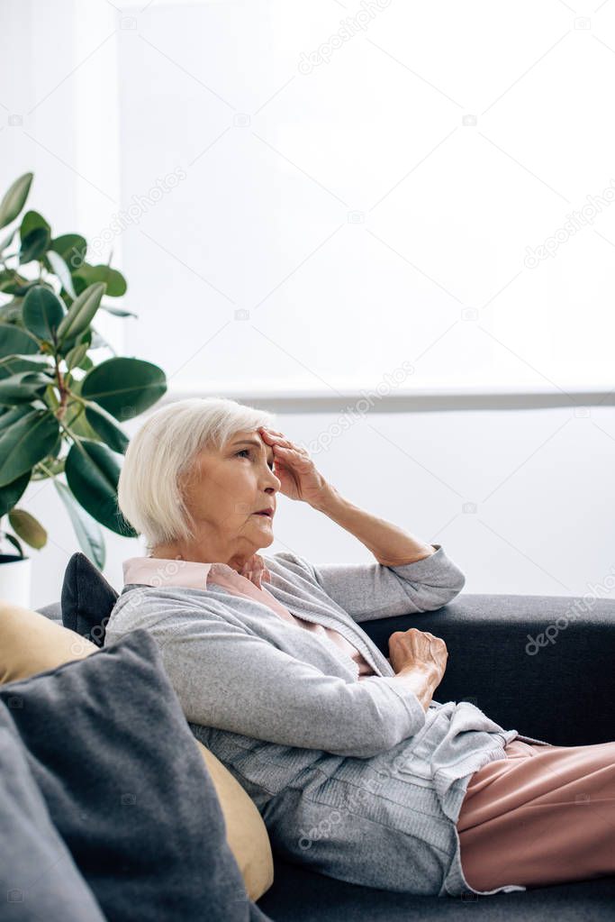 Senior woman having headache and sitting on sofa in apartment