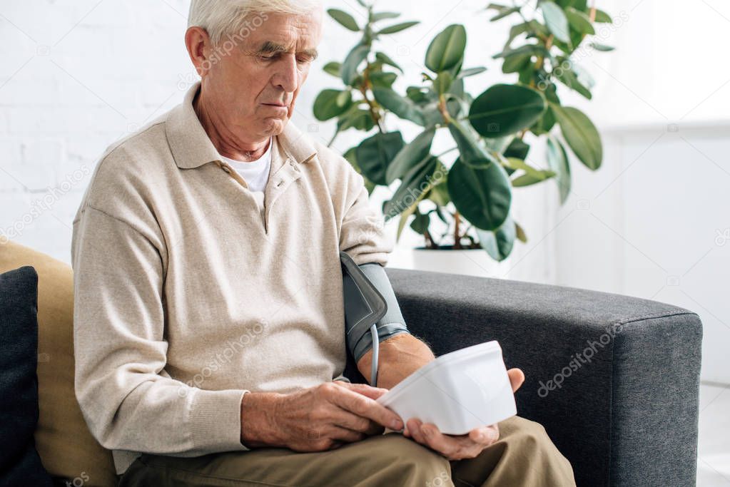 senior man measuring blood pressure and sitting on sofa in apartment 