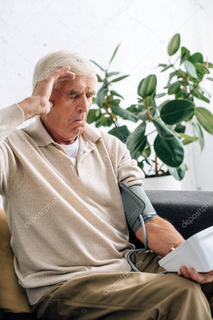 shocked senior man measuring blood pressure and sitting on sofa in apartment 