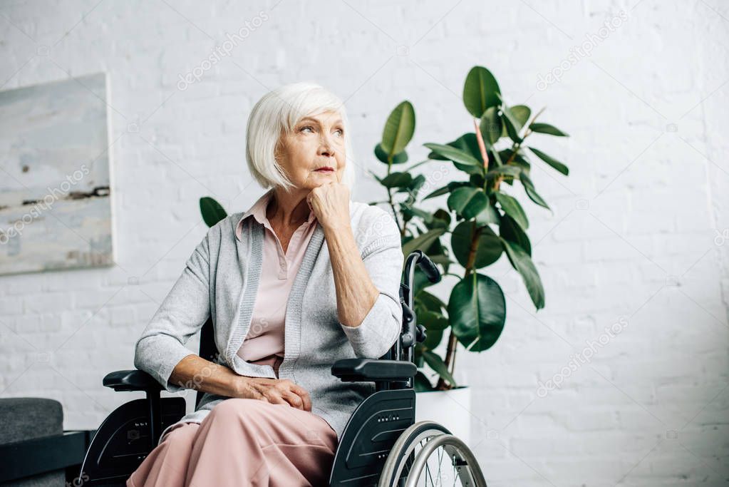 dreamy senior woman sitting in wheelchair and looking away in apartment 