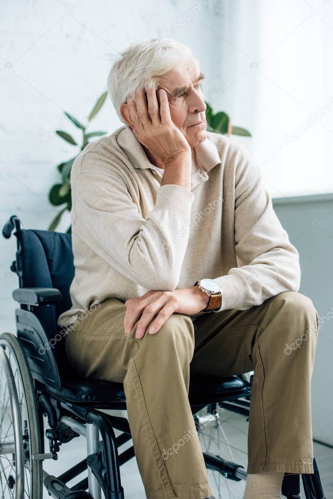 handsome and dreamy senior man sitting in wheelchair in apartment 