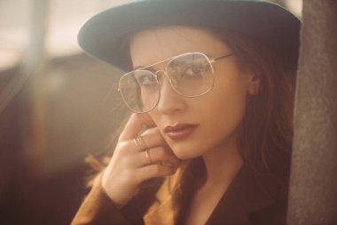 fashionable woman posing in hat, eyeglasses and brown jacket on roof clipart