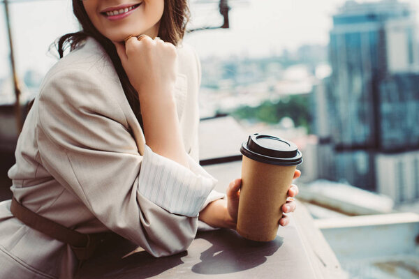 cropped view of smiling girl in beige suit posing on roof with coffee to go