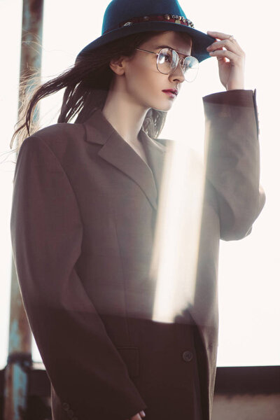 elegant fashionable woman posing in hat, eyeglasses and brown jacket on roof