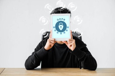Bilgisayar korsanı masada oturuyor ve elinde GDPR illüstrasyonlu dijital tablet tutuyor
