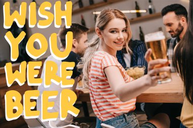selective focus of smiling young woman looking at camera while holding glass of light beer near wish you were beer illustration clipart