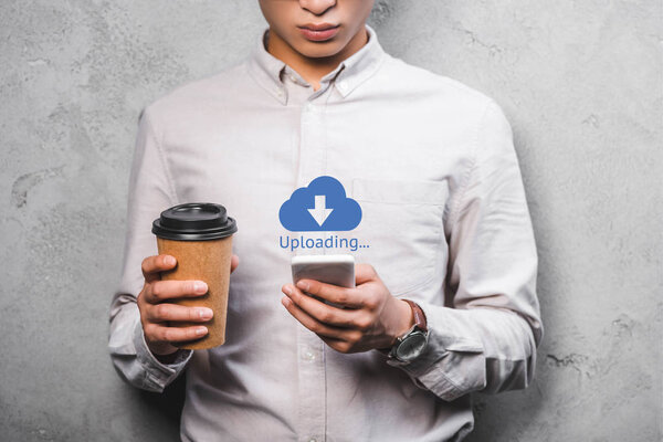 cropped view of seo manager holding paper cup, using smartphone and standing near uploading illustration