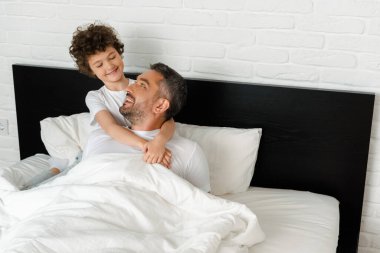 Kıvırcık çocuk yatak odasında neşeli babasına sarılıyor. 