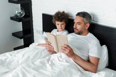 Sakallı baba yatak odasında kıvırcık oğlunun yanında kitap okuyor. 