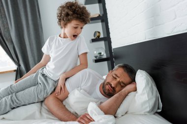 curly boy screaming and looking at bearded father lying on bed  clipart