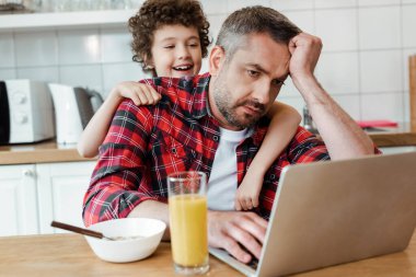 selective focus of happy son touching tired freelancer father working from home clipart
