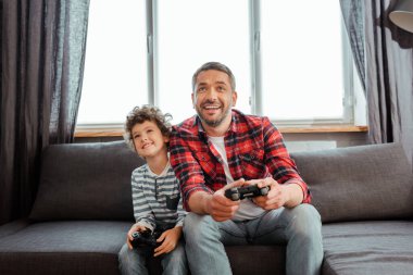 KYIV, UKRAINE - 14 Mayıs 2020: oturma odasında video oyunu oynayan yakışıklı baba ve mutlu oğul 