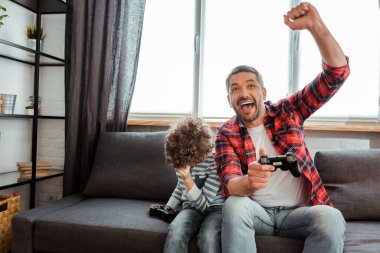 KYIV, UKRAINE - MAY 14, 2020: happy father celebrating triumph near curly son while holding gamepad in living room  clipart