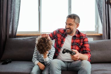 KYIV, UKRAINE - 14 Mayıs 2020: mutlu baba oturma odasında video oyunu oynarken üzgün ve kıvırcık oğluna dokunuyor 
