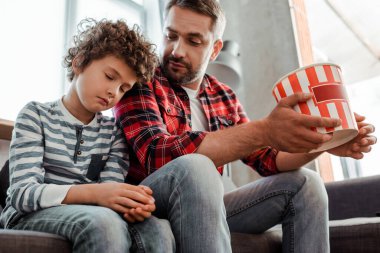 Baba elinde patlamış mısır kovası, uykulu oğlana bakıyor. 