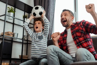 Heyecanlı çocuk, babasıyla şampiyonluk maçı izlerken elinde futbol tutuyor.