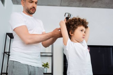 Yakışıklı baba halterlere dokunurken kıvırcık oğlan evde egzersiz yapıyor.