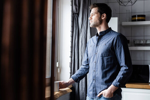 sad man holding cup of coffee while looking through window at home on quarantine