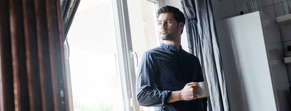 handsome man holding cup of coffee and standing near window at home on quarantine, horizontal concept