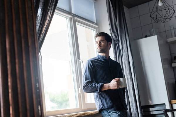 handsome pensive man holding cup of coffee and standing near window at home on self isolation