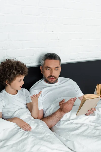 ベッドルームで可愛い息子のそばで読書する髭の父親 — ストック写真