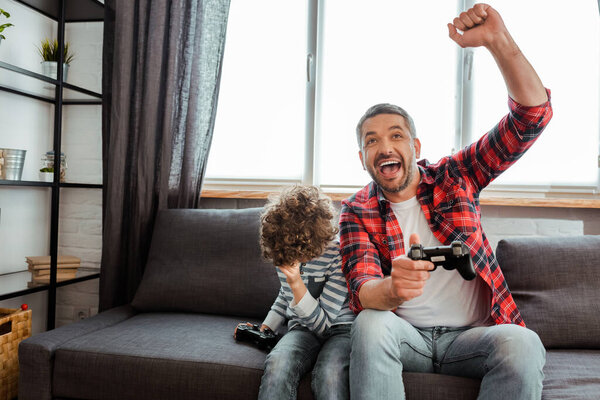 KYIV, UKRAINE - MAY 14, 2020: happy father celebrating triumph near curly son while holding gamepad in living room 