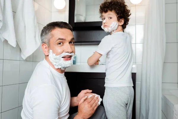 洗澡间靠近卷曲儿子的相机前 有选择性地聚焦有剃须泡沫的父亲的脸 — 图库照片