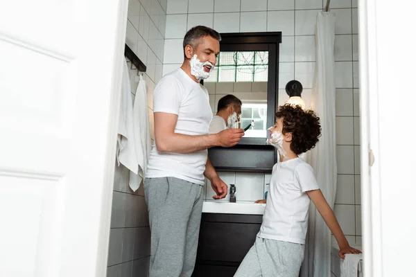 可愛い息子の近くに剃刀を持って顔にシェービングフォームを持つハンサムなお父さん — ストック写真
