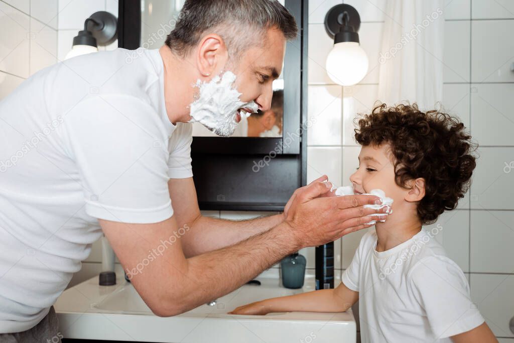 side view of happy father applying shaving foam on face of cute son in bathroom 