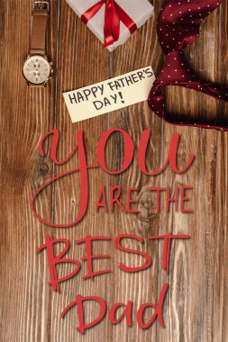 Top view of gift box with red bow, greeting card with lettering happy fathers day and mens red tie and wristwatch on wooden background, you are the best dad illustration clipart