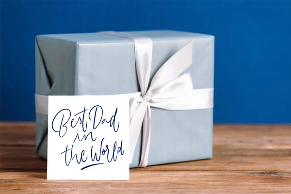 selective focus of greeting card with best dad in the world illustration and gift box with white bow isolated on blue