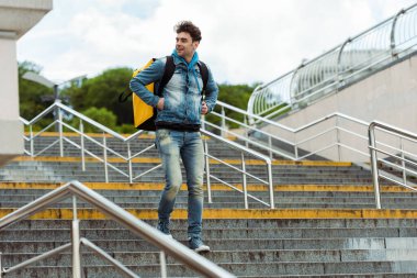 Selective focus of smiling courier with thermo backpack walking down on stairs with railing  clipart