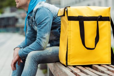 Şehir sokağındaki bankta termal çantanın yanında oturan kurye görüntüsü. 