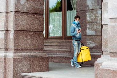 Binanın yakınında akıllı telefon kullanan tıbbi maskeli bir teslimatçı. 