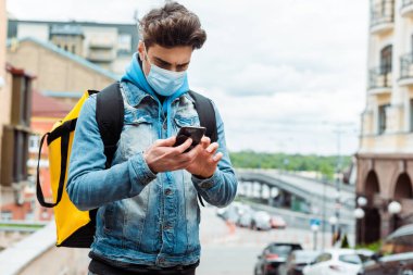 Şehir caddesinde akıllı telefon kullanan tıbbi maskeli bir kurye. 