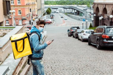 Tıbbi maskeli kuryenin şehir sokaklarında akıllı telefon kullanarak termo sırt çantalı yan görüntüsü 